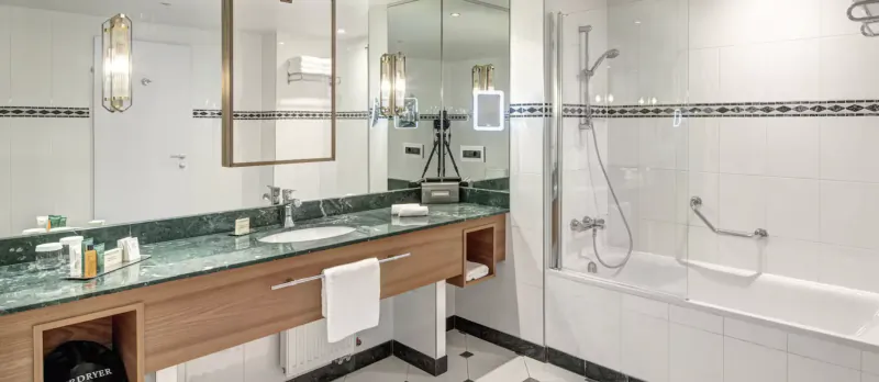 Modern hotel bathroom with marble vanity, double sinks, large mirror, and white bathtub with glass shower