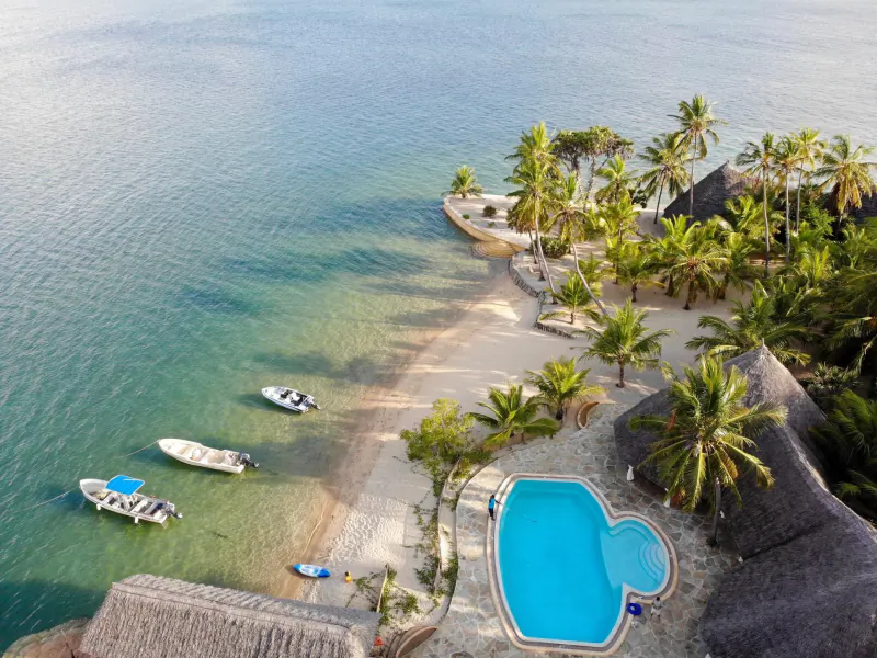 Aerial view of Manda Bay Lodge in Lamu, Kenya, showing a heart-shaped pool, thatched-roof villas, palm trees, and boats along a sandy beach