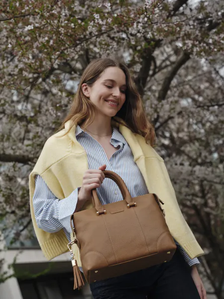 Smiling woman in yellow knit vest and striped shirt holding a brown leather handbag under blooming cherry blossoms