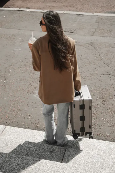 Woman with long dark hair in brown blazer and jeans, carrying silver suitcase and iced coffee, walking up stone steps