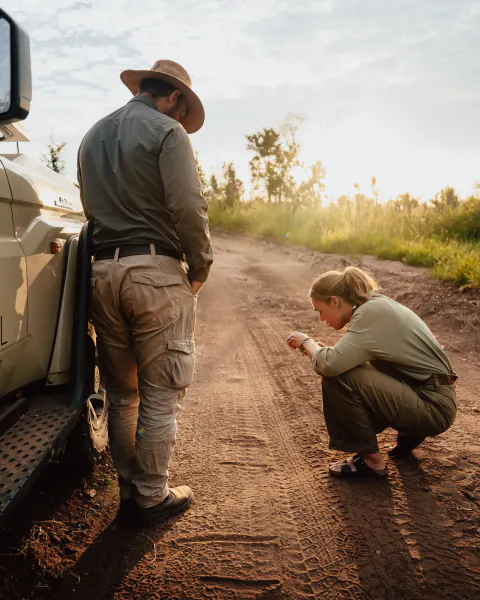 Man in safari hat and woman in green shirt examine dirt road tracks beside a safari vehicle at sunset