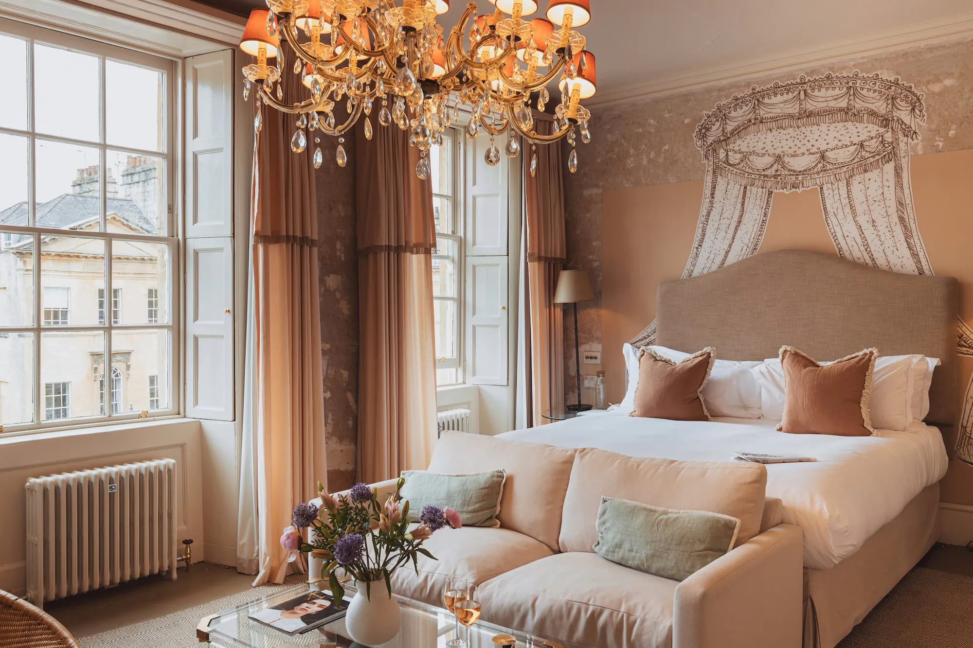 Elegant hotel bedroom with large bed, crystal chandelier, sofa, and tall windows overlooking historic buildings
