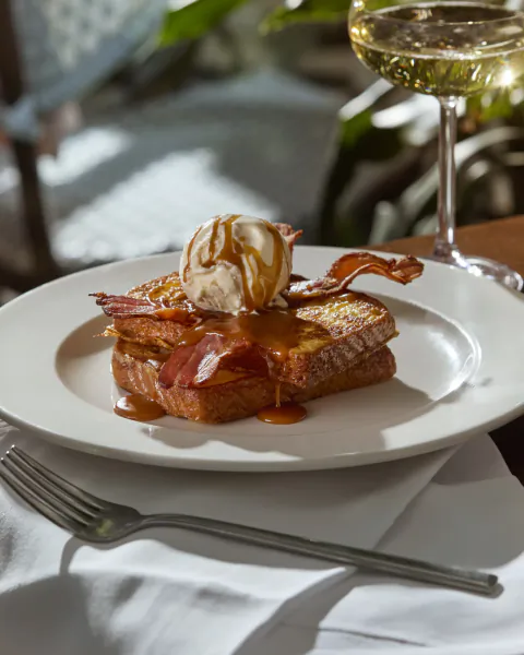 French toast topped with bacon, ice cream, and caramel sauce on a white plate beside a glass of white wine