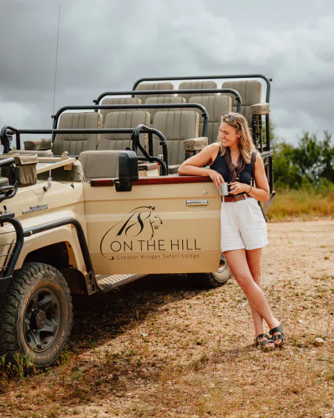 Woman leaning on safari vehicle with 'ON THE HILL Greater Kruger Safari Lodge' logo, holding a mug in savanna setting