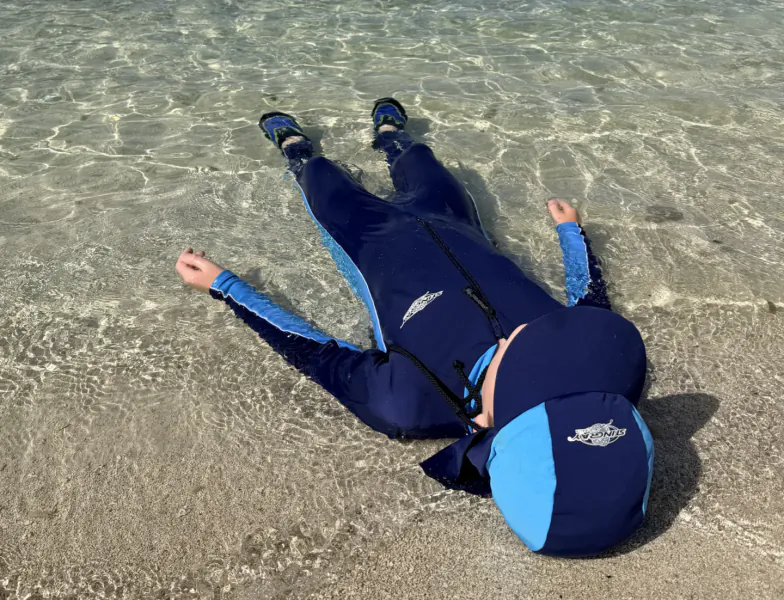 Child in blue and black full-body sun protection swimsuit and cap lying on wet sand in shallow clear water