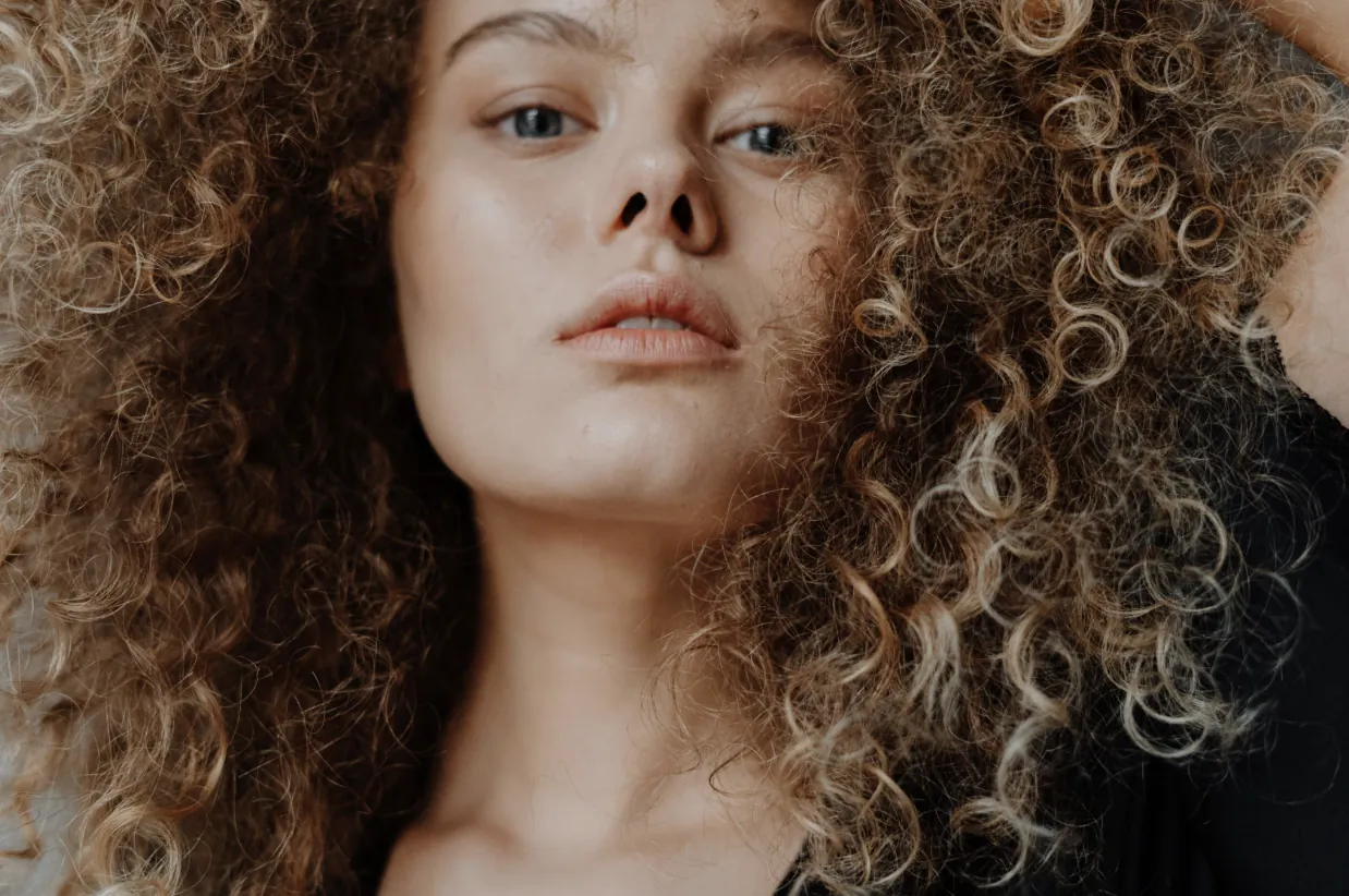 Close-up of a woman with voluminous curly brown and blonde hair, neutral expression, looking at camera