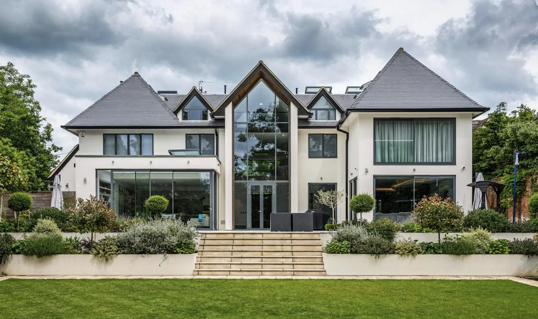 Modern luxury house with large glass windows, gabled roof, and landscaped front garden under cloudy sky