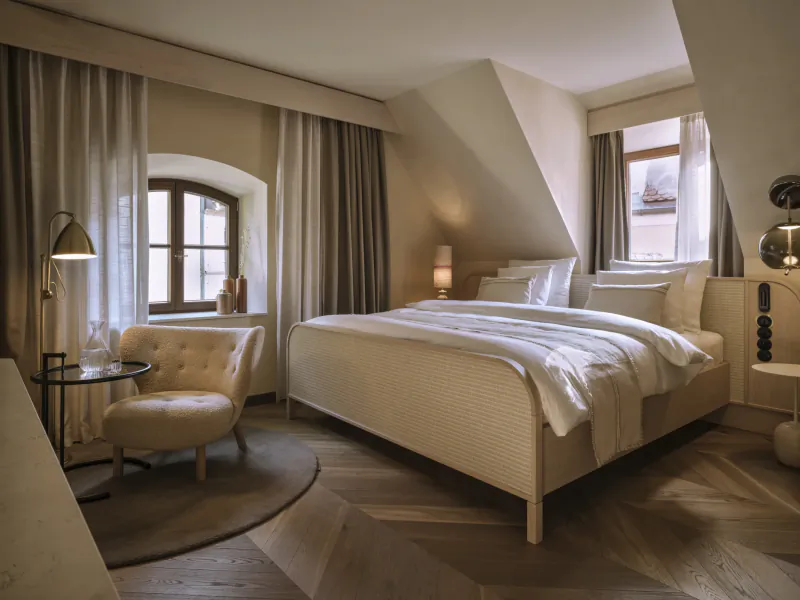 Bright hotel bedroom with a large bed, beige armchair, arched window, and warm lighting at Platzl Hotel Munich
