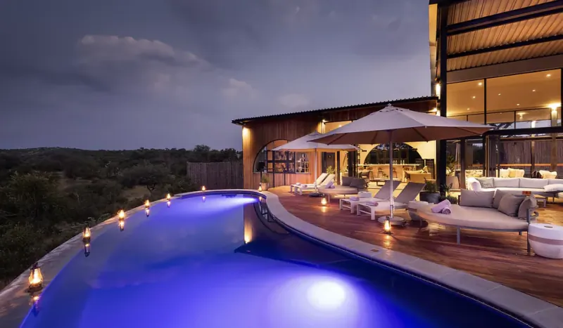 Luxury safari villa at dusk with illuminated infinity pool, lounge chairs, umbrellas, and modern glass-and-wood architecture overlooking the African bushveld