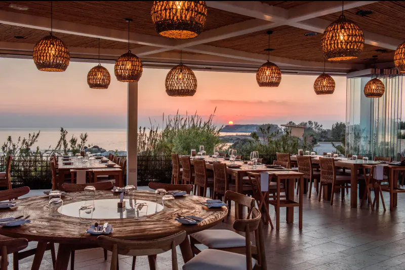 Elegant open-air restaurant at sunset with round wooden tables, woven pendant lights, and ocean view overlooking the sea