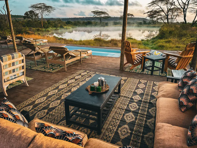 Luxury safari lodge deck with lounge chairs, sofas, and a pool overlooking a misty lake and savanna at sunrise