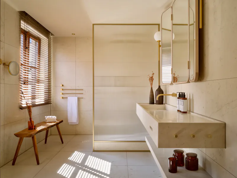 Modern minimalist bathroom with marble walls, floating vanity, gold-framed shower, wooden bench, and sunlight through blinds