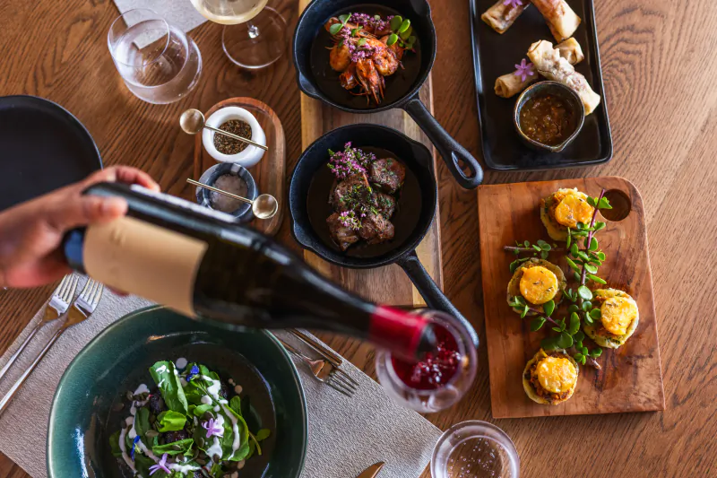 Hand pouring red wine over a table of gourmet dishes including steak, shrimp, and appetizers on a wooden table