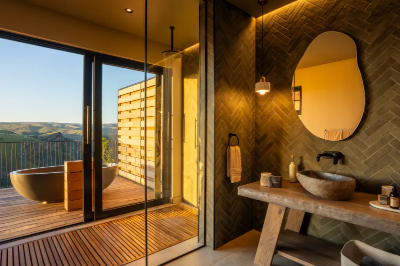 Modern bathroom with stone sink, round mirror, and open glass doors to a wooden deck with freestanding tub overlooking hills