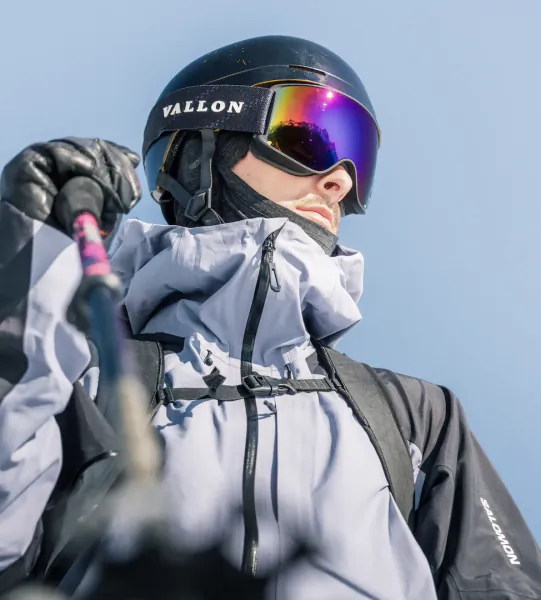 Close-up of skier in black VALLON helmet with iridescent goggles, light blue jacket, holding ski pole against clear blue sky