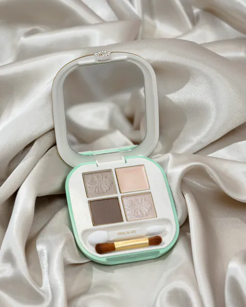Open Paul & Joe eyeshadow palette with four floral-embossed shades, mirror, and dual-ended brush on white satin fabric