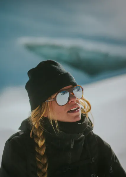 Woman in black winter jacket and beanie wearing reflective sunglasses, standing in snowy landscape with braided blonde hair