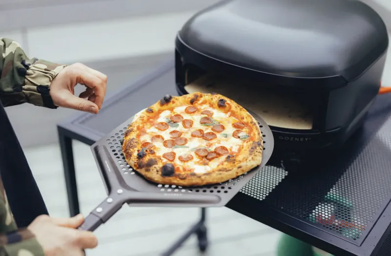 Person holding pepperoni pizza on peel in front of black Gozney Arc Lite pizza oven