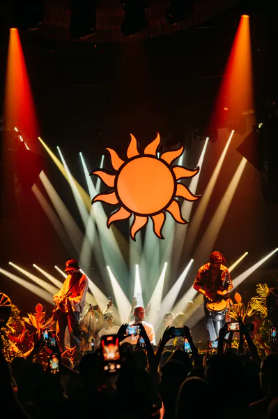 Concert stage with sun logo backdrop, two guitarists performing under red spotlights, crowd filming with phones