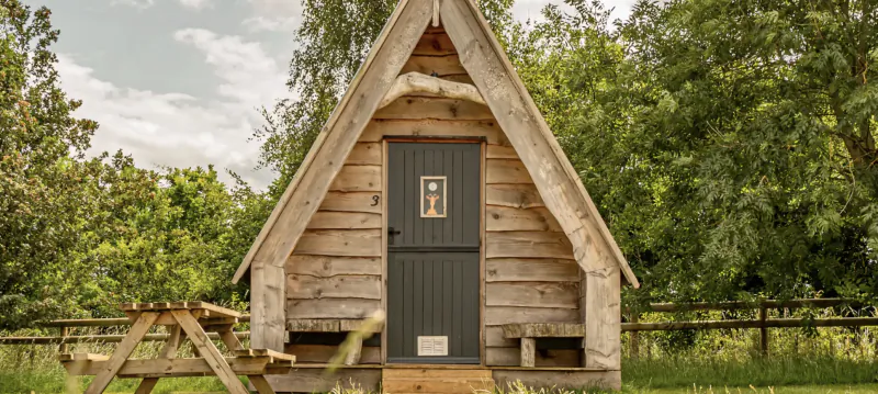 Wooden A-frame glamping cabin with dark grey door and picnic table in a grassy field surrounded by trees