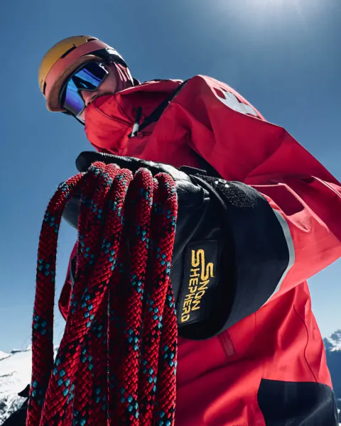 Snowboarder in red jacket holding coiled red and blue climbing rope with SnowShepherd logo on black pouch