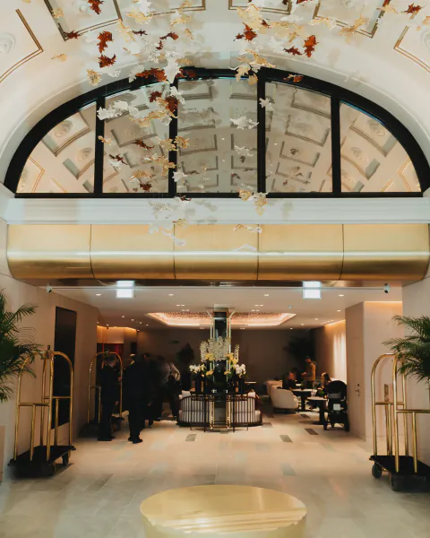 Elegant hotel lobby with gold reception desk, arched windows, hanging floral installation, and guests near brass luggage carts