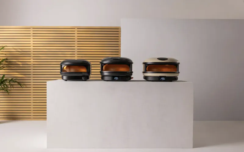 Three compact Gozney Arc Lite pizza ovens in black, black, and beige lined up on a white pedestal against a wooden slat wall