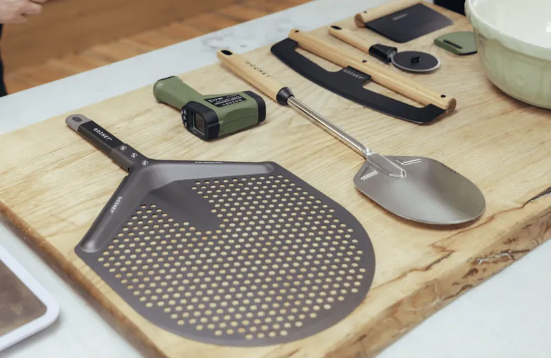 Gozney pizza peel with perforated surface and metal scoop on wooden cutting board with kitchen tools