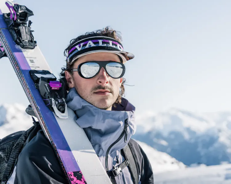 Man in ski gear holding skis on snowy mountain, wearing VALLON visor and reflective goggles