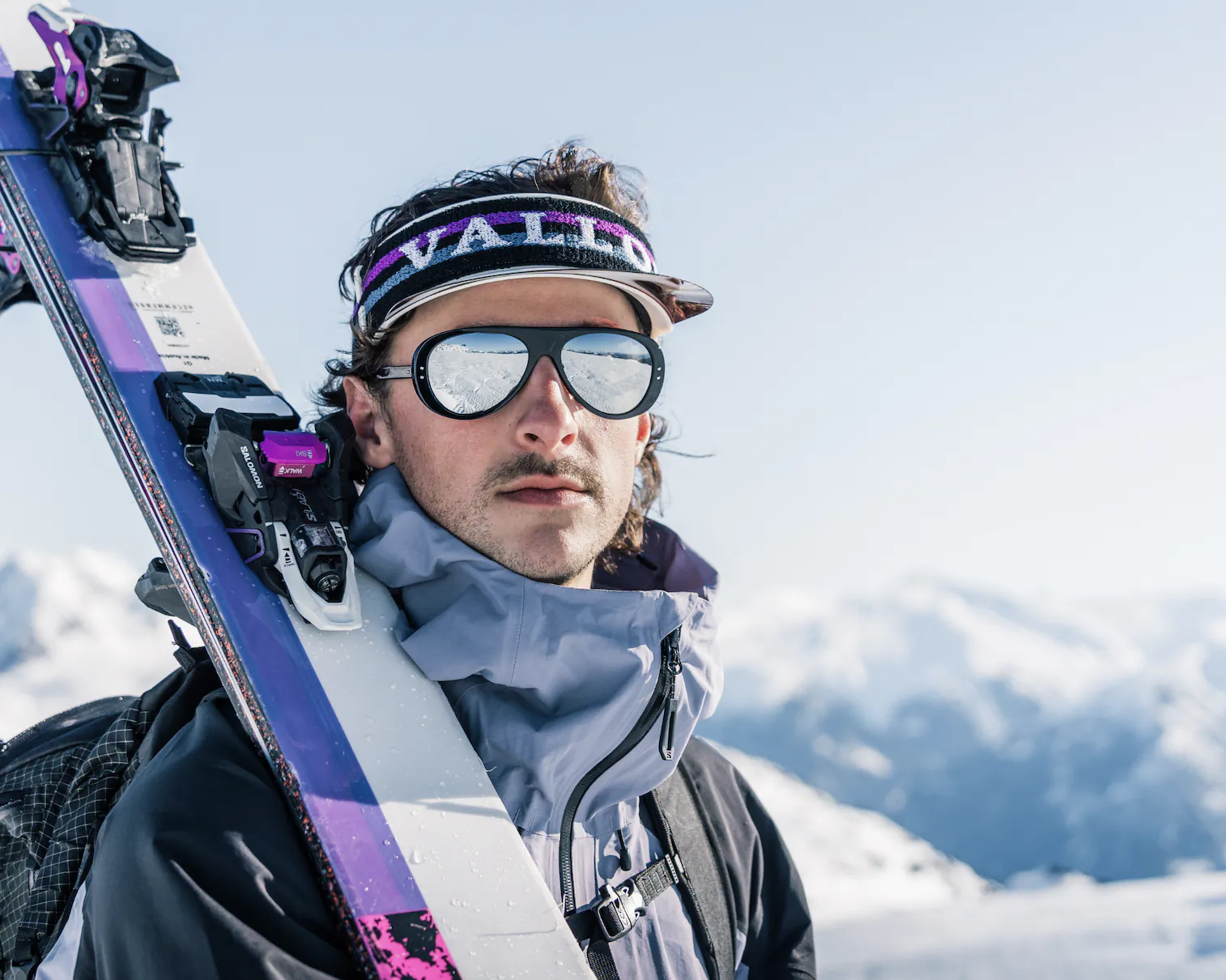 Man in ski gear holding skis on snowy mountain, wearing VALLON visor and reflective goggles