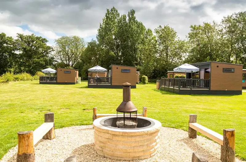 Luxury glamping pods with decks and umbrellas in a grassy field, stone fire pit in foreground