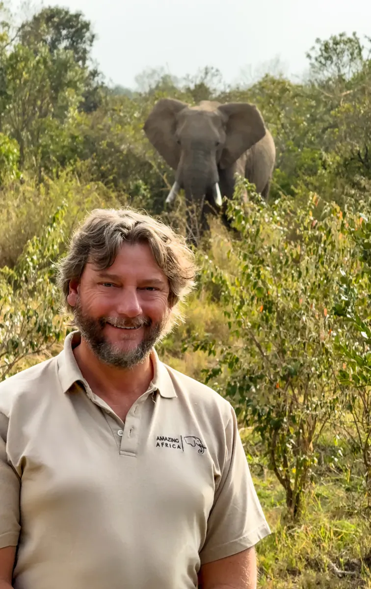 Smiling man in beige polo shirt with Amazing Africa logo stands in front of an elephant in the African bush