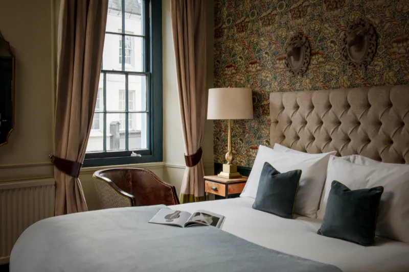 Luxury hotel bedroom with tufted beige headboard, white bedding, open magazine on bed, and patterned wallpaper