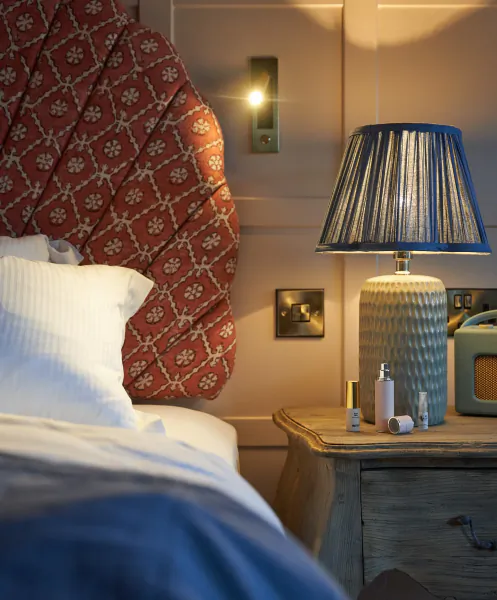 Cozy bedroom with red patterned headboard, white bedding, lit blue lamp on wooden nightstand with toiletries and vintage radio