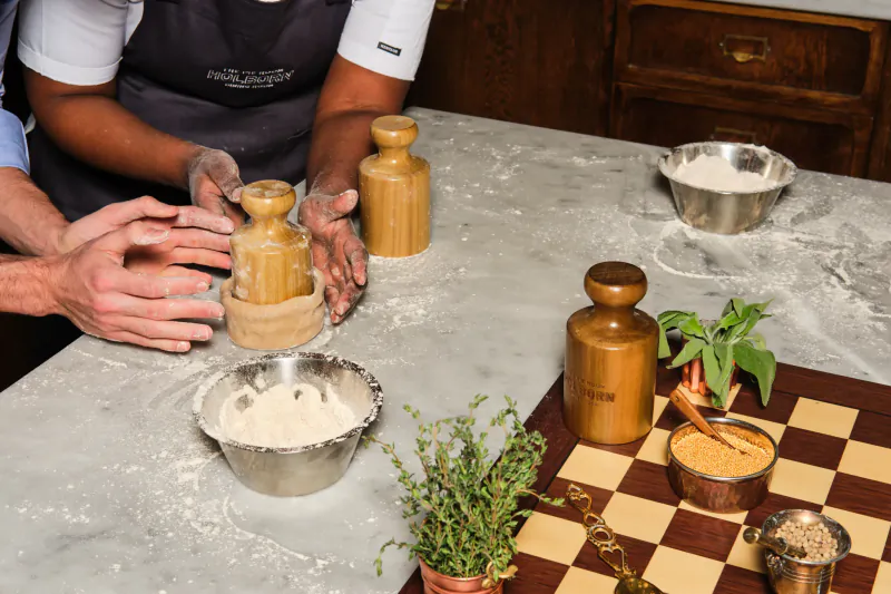 Chef in apron rolling dough with wooden rolling pin on marble counter, surrounded by flour bowls, herbs, and spices