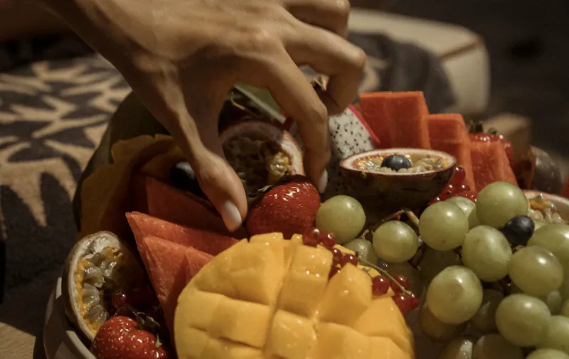 Hand reaching into a vibrant fruit platter with mango, strawberries, grapes, watermelon, and passion fruit at a beachside setting