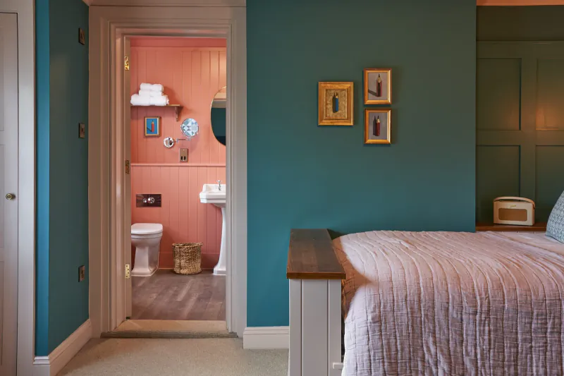 Bedroom with teal walls and pink bed, open doorway showing pink en-suite bathroom with toilet and sink