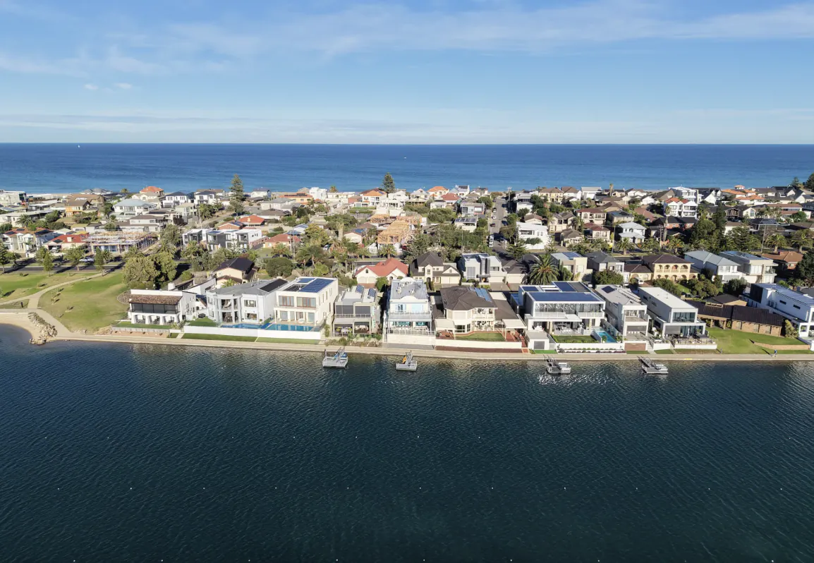 Aerial view of modern waterfront homes along a calm Australian coastline with boats docked in front