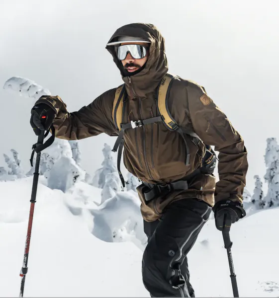 Man in brown hooded winter jacket and ski goggles holding poles on snowy mountain slope