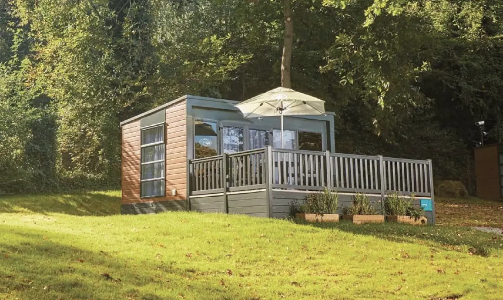 Wooden cabin with deck, umbrella and planters on a grassy hill surrounded by trees