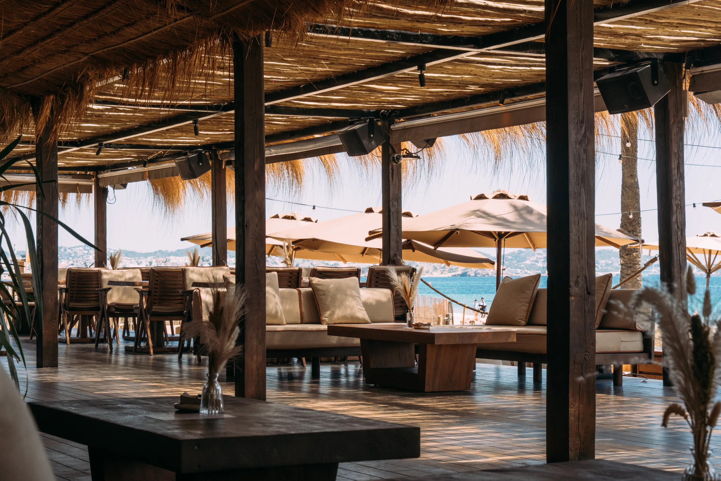 Beachouse Ibiza: open-air beachfront lounge with wooden tables, sofas, thatched roof, and ocean view