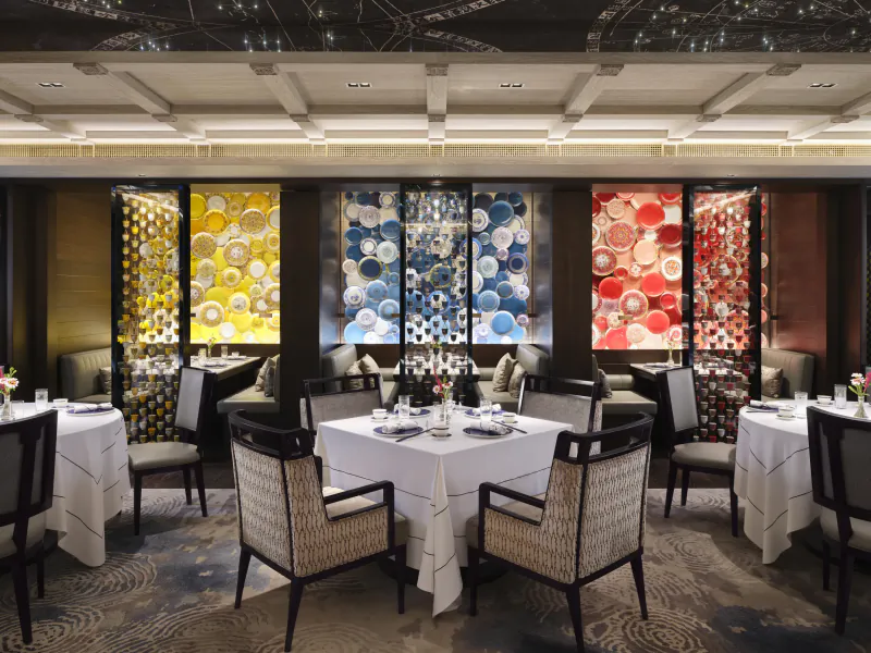 Elegant fine dining restaurant with white tablecloths, patterned chairs, and colorful circular mosaic wall panels in yellow, blue, and red.