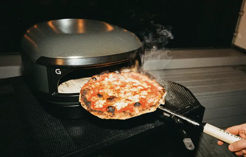 Hand pulling a charred pepperoni pizza from a Gozney Arc Lite pizza oven with steam rising