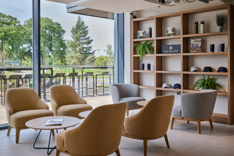 Modern hotel lounge with mustard and grey armchairs around a round table, large window overlooking trees, and wooden shelving unit displaying The Belfry sign and caps