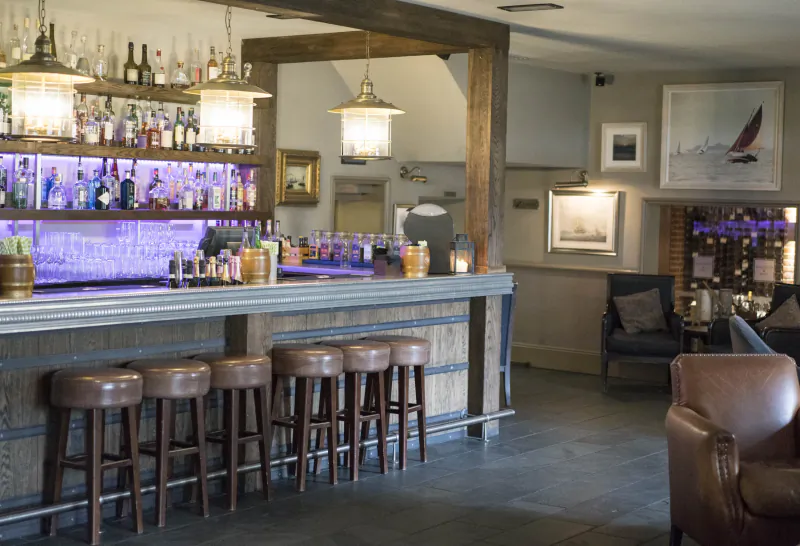 Hotel bar with wooden counter, five brown leather stools, backlit liquor shelves, and leather armchairs in a lounge area