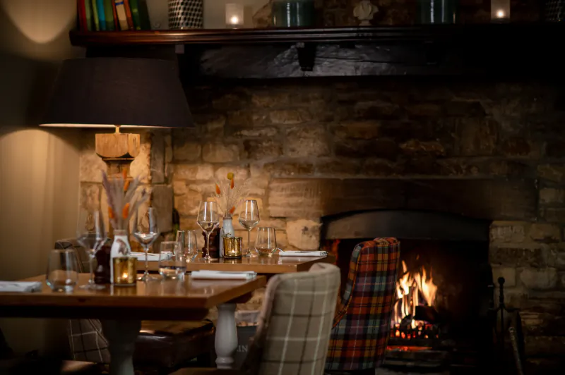 Cosy restaurant table set for dinner beside a lit stone fireplace with wine glasses and a plaid chair