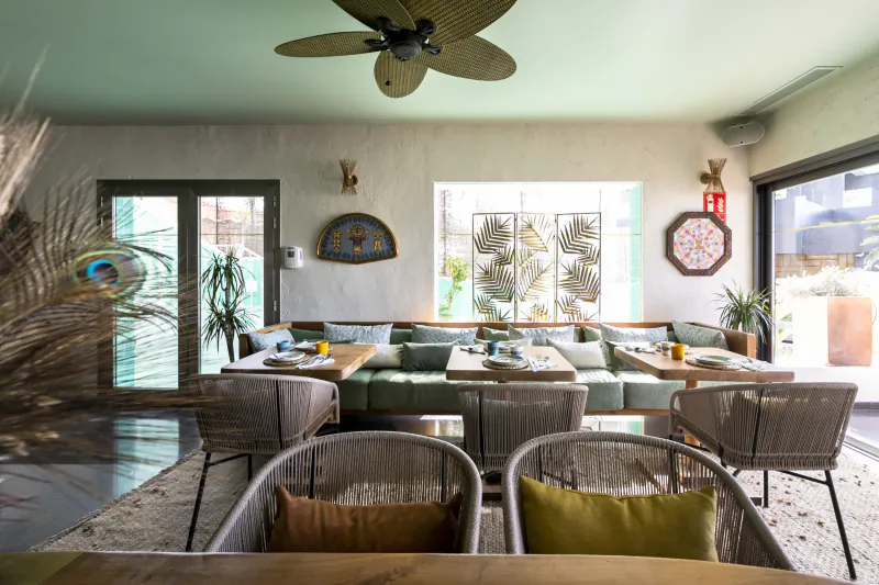 Modern Ibiza-style lounge with green sectional sofa, woven chairs, wooden tables, and tropical wall decor