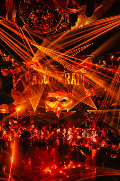 Crowded nightclub with orange laser beams, hanging masks, and glowing 'MASQUERADE' sign above a party crowd