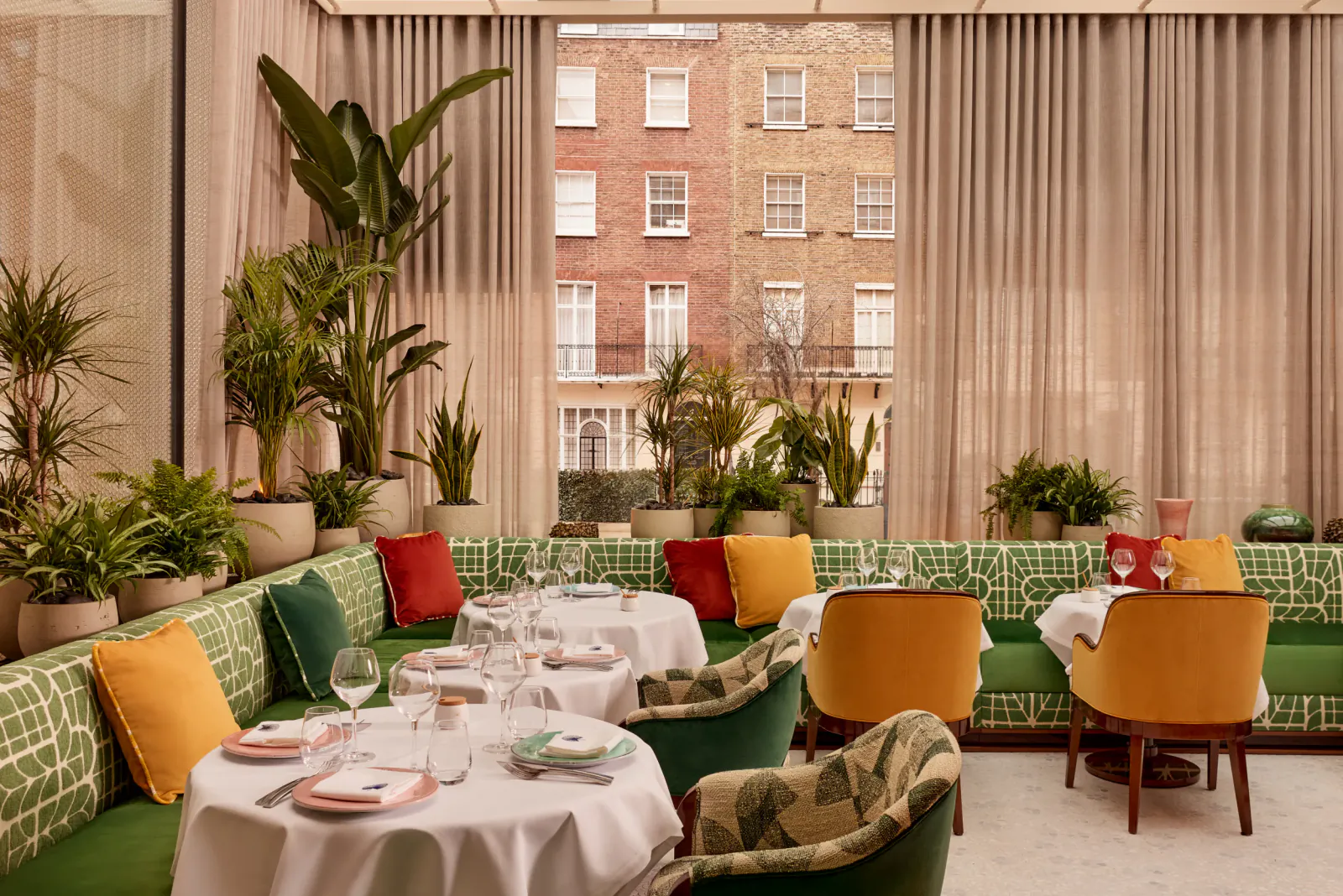 Elegant restaurant interior with green patterned banquettes, colorful cushions, round tables set with white tablecloths, and large windows overlooking a brick building