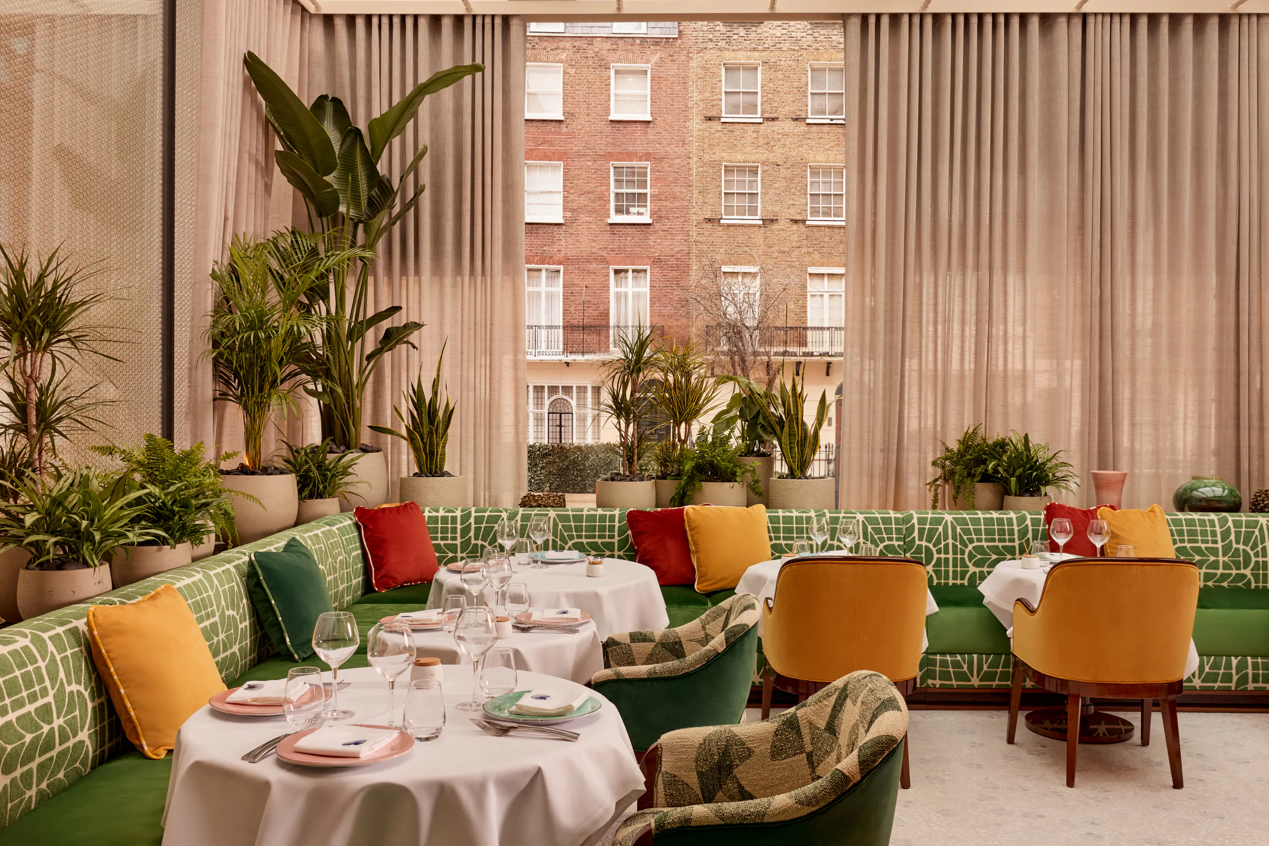 Elegant restaurant interior with green patterned banquettes, colorful cushions, round tables set with white tablecloths, and large windows overlooking a brick building